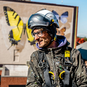A artist wearing a helmet and harness stands outdoors near a vibrant mural featuring large yellow and black butterflies. The setting appears urban, with buildings and clear skies visible in the background. The worker's gear includes safety straps and tools, emphasizing a professional work environment.