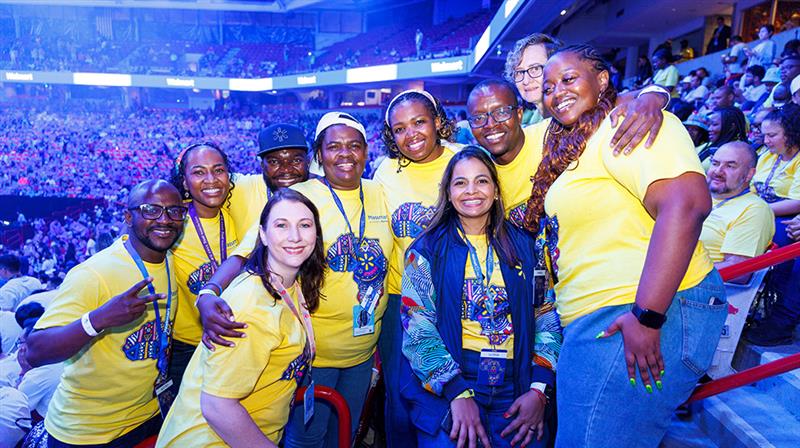 A group of individuals wearing bright yellow shirts with colorful designs poses together in a crowded indoor arena. The setting features a vibrant atmosphere with a large audience in the background, illuminated by blue lighting.