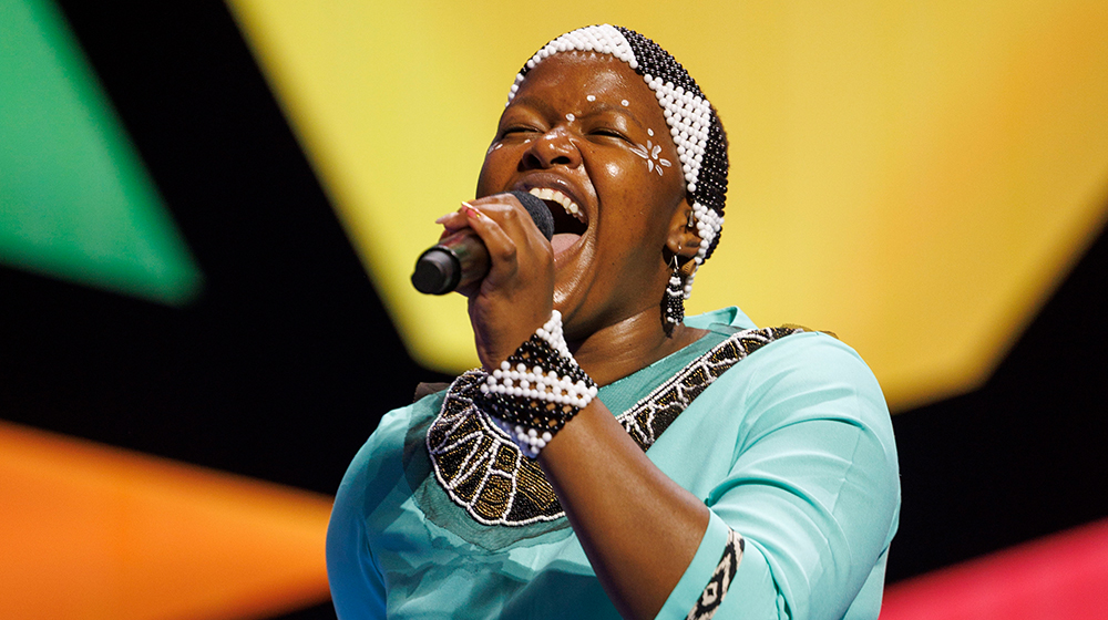 A performer wearing intricate beaded accessories and a turquoise outfit is captured mid-performance. The setting features a vibrant, colorful backdrop with geometric patterns. The individual holds a microphone, suggesting a live event or concert.