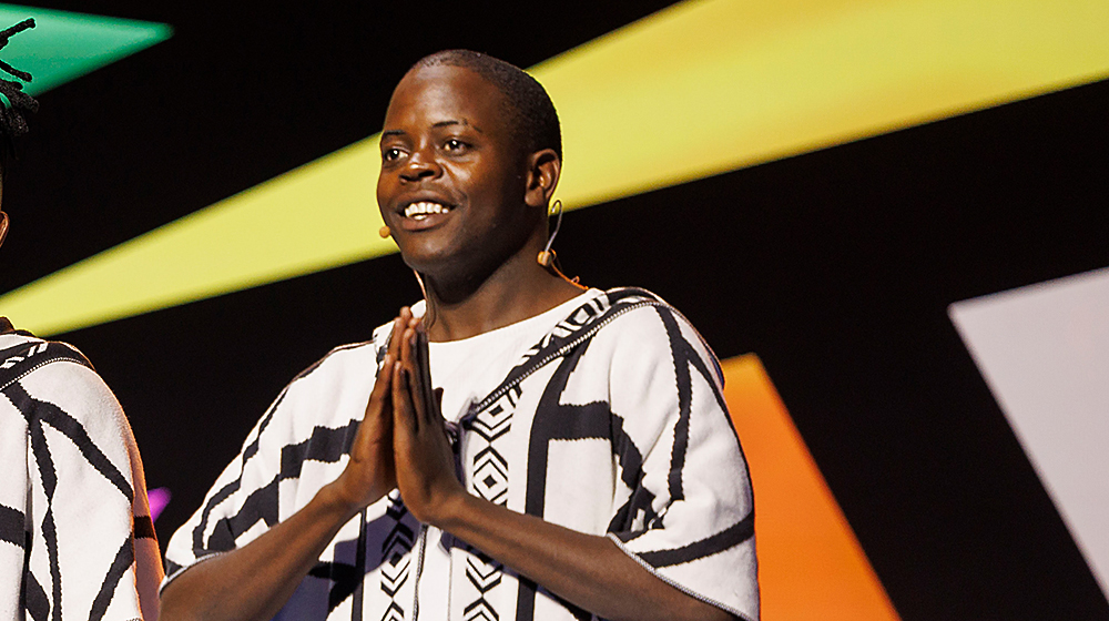A person wearing a black-and-white patterned garment stands on a stage with hands clasped together. The background features vibrant geometric shapes in yellow, orange, and white. 
