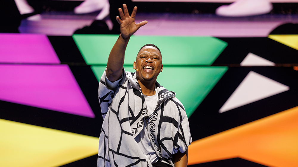 A person wearing a patterned outfit stands on a stage with a colorful geometric background. The setting features bold shapes in green, yellow, purple, and white. The individual raises one hand in a dynamic gesture, creating a lively and engaging atmosphere.