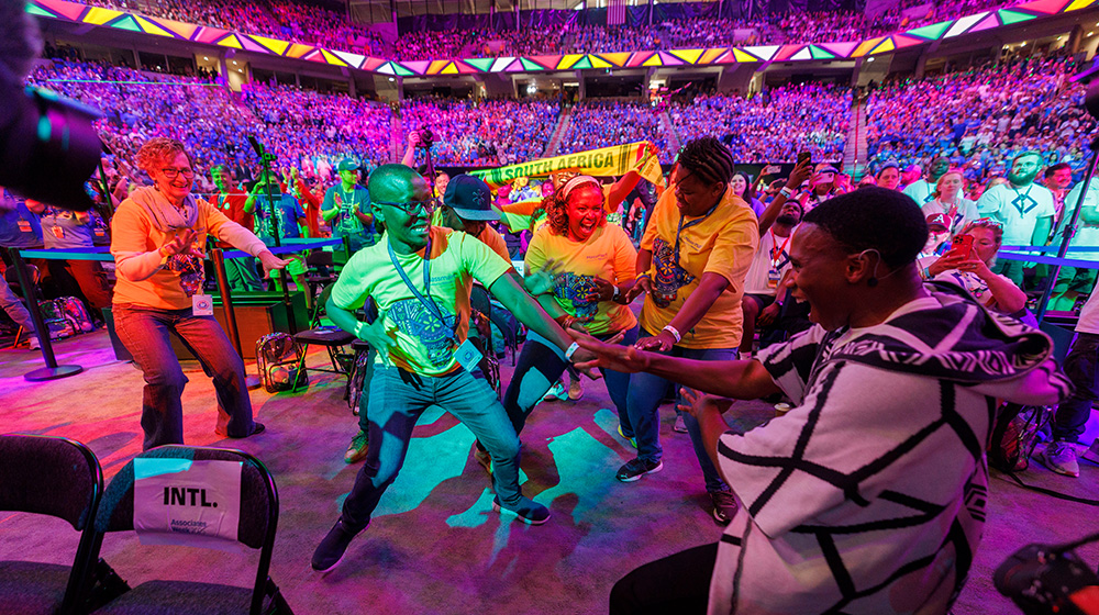 brant indoor event featuring a group of people engaged in dancing. The setting is a large, colorful arena with bright lighting and a lively atmosphere. Participants are wearing yellow and other colorful clothing, with a visible banner in the background. The scene captures energy and community spirit.