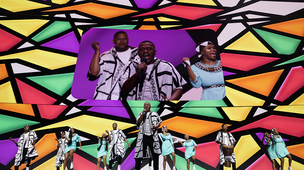 A group of performers dressed in traditional black-and-white patterned attire and turquoise outfits perform on a brightly lit stage. The background features a vivid geometric design with bold colors such as red, yellow, green, and purple. 