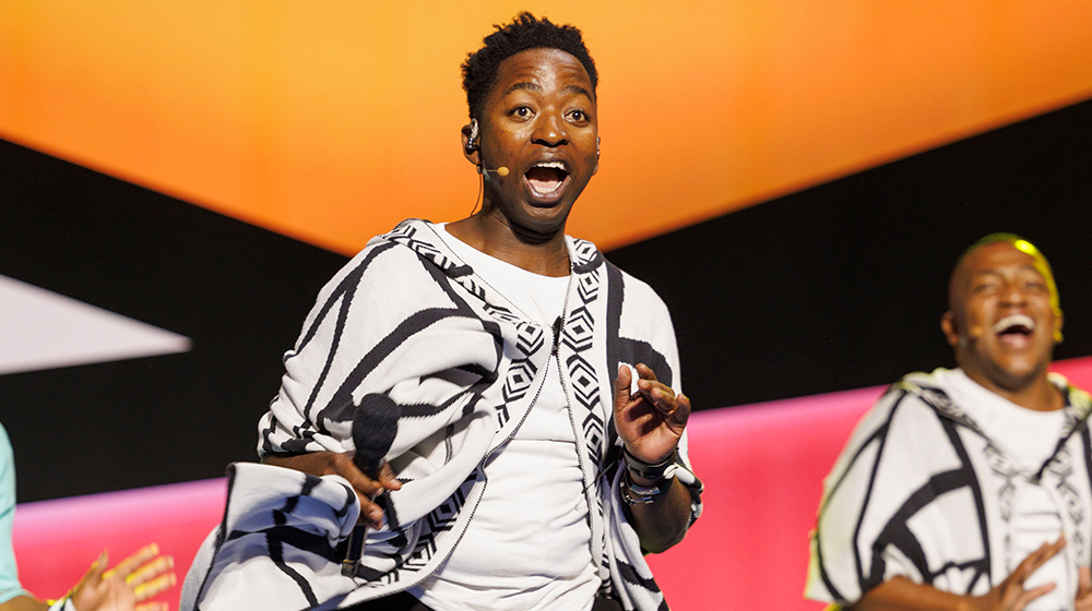 A performer wearing a black-and-white patterned shawl over a white shirt is captured mid-motion on stage. The setting features vibrant geometric backdrops in orange, pink, and black tones. The scene suggests a lively performance or event with additional performers partially visible.