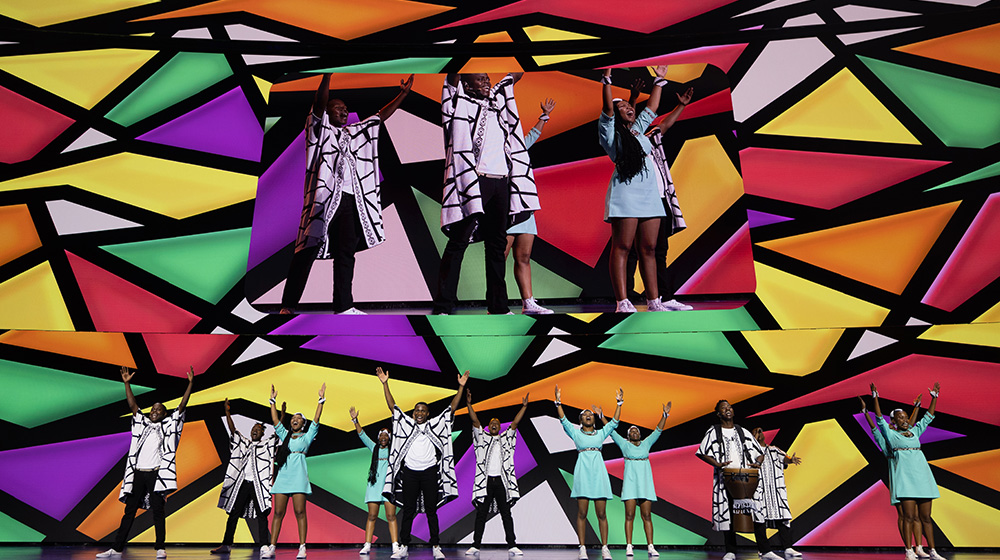 A dynamic group performance on a brightly lit stage featuring bold geometric patterns in vivid colors. Performers are dressed in contrasting black-and-white outfits and pastel blue dresses, creating a striking visual contrast. The scene exudes energy and creativity, with a diverse group of individuals engaging in synchronized movements. The backdrop features a kaleidoscope of colors, enhancing the lively atmosphere.