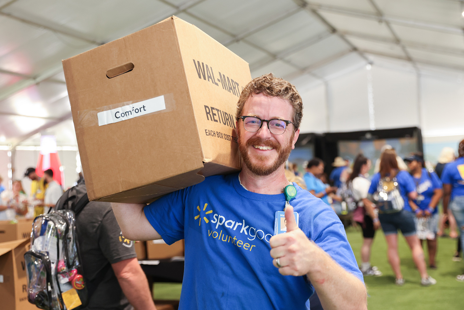 A photo of Walmart associate volunteering and wearing Spark good t-shirt, carrying a large box on his shoulder possibly containing donated items from Walmart. He is smiling and showing thumbs up. 